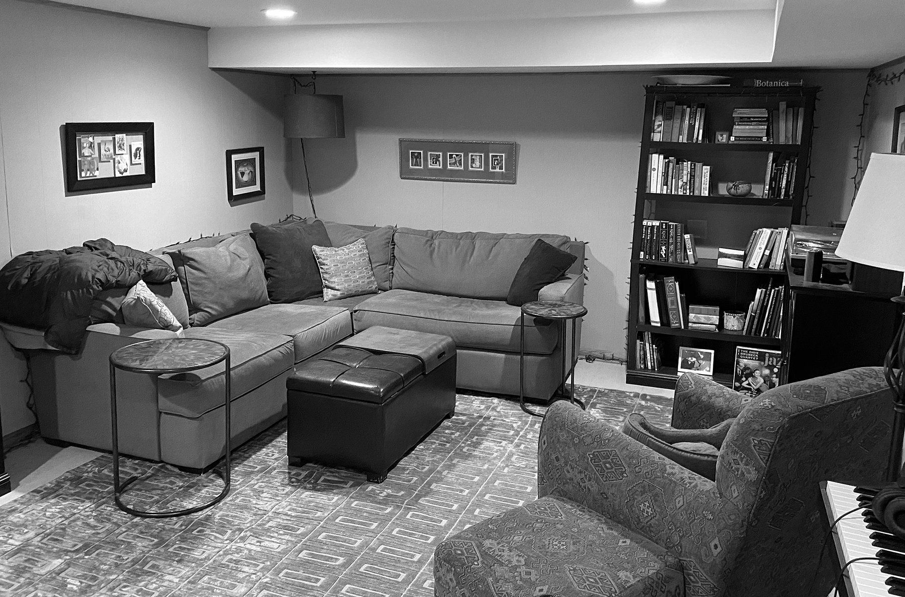 A black and white image of a basement entertaining space with a couch, recliner and bookcase.